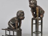 two bronze statues of children sitting on stools on a white surface