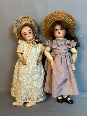 une paire de poupées anciennes, coiffées et vêtues, exposées sur un fond gris