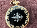 une montre de poche ancienne, noire et dorée, posée sur un tissu rose