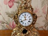 une horloge en or posée sur une table en bois, avec un papier peint à motifs floraux en arrière-plan