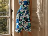 La femme se tient debout devant une fenêtre, vêtue d'une robe à fleurs bleues et blanches.