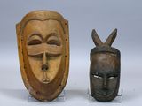 Deux masques en bois sont posés sur un socle, sur une surface blanche.