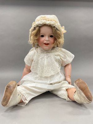 une poupée ancienne assise par terre, vêtue d'une robe blanche et d'un chapeau