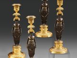 quatre bougeoirs en bronze ornés de figurines, posés sur des socles, sur un fond gris