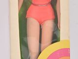 une poupée Barbie dans une boîte, posée sur une surface blanche, vêtue d'une robe rose