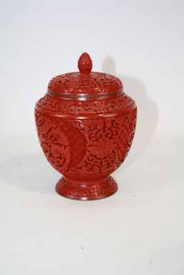 un vase rouge muni d'un couvercle, posé sur une surface blanche