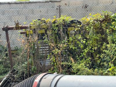 a large pipe that is next to a fence with plants growing out of it