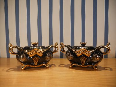 une paire de théières noires et dorées posées sur une table en bois, devant un papier peint à rayures bleues et blanches