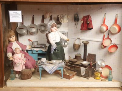 deux poupées dans une vitrine, avec des casseroles et des poêles accrochées au mur