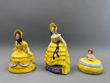 trois figurines représentant des femmes en robe jaune et chapeau, posées sur un fond gris