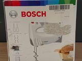 Mixeur plongeant Bosch dans son emballage, posé sur une table en bois avec un arrière-plan multicolore