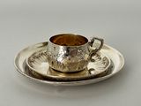 une tasse et une soucoupe anciennes en argent, posées sur une soucoupe, sur une surface blanche
