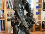 bronze statue of a man sitting on a stool with a bookcase in the background