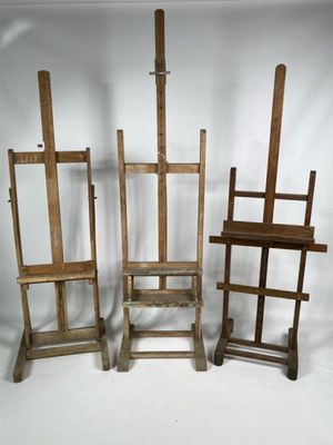 Trois chevalets en bois et une chaise sont disposés devant un fond blanc.