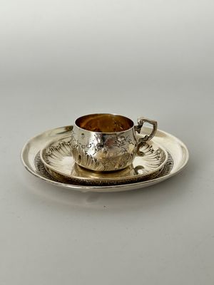 une tasse et une soucoupe anciennes en argent, posées sur une soucoupe, sur une surface blanche