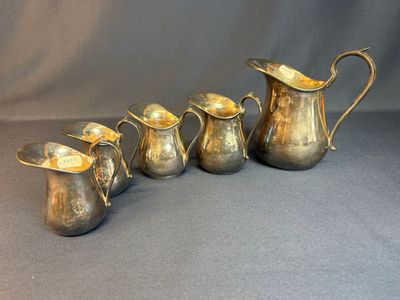 five antique silverplated jugs and a pitcher on a grey background