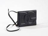 Le petit sac noir est posé sur une surface blanche et est muni d'une bandoulière noire.