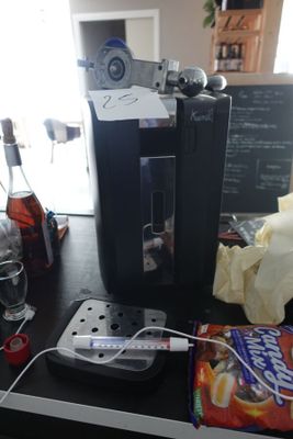 une cafetière posée sur une table, à côté d'une bouteille de soda et d'un paquet de chips