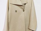 manteau en laine beige suspendu à un cintre devant un mur blanc, sur fond blanc