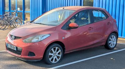 une voiture rouge garée sur un parking, avec des vélos en arrière-plan