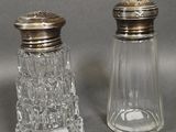 deux salières et poivrières en verre avec des couvercles argentés, posées sur un plan de travail gris