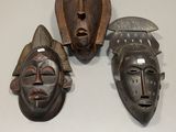 trois masques en bois posés sur une surface blanche, dont l'un porte un casque
