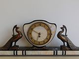 une horloge posée sur une étagère, surmontée de deux oiseaux