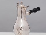 une cafetière ancienne en argent, avec une poignée noire, posée sur une table blanche