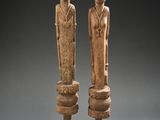 paire de figurines en bois sculpté posées sur un socle, sur fond gris