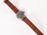 La montre repose sur une surface blanche et est dotée d'un bracelet marron.