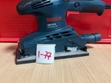 une ponceuse orbitale Bosch posée sur une table, à côté d'une boîte rouge