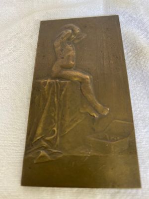 plaque en laiton représentant une femme assise sur une table, les jambes croisées