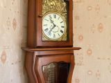 une horloge de parquet placée dans un coin de la pièce, avec derrière elle un mur recouvert de papier peint