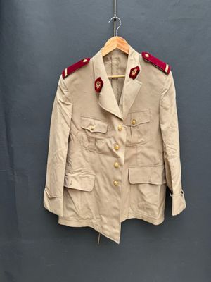 une veste beige aux bordures rouges, suspendue à un cintre devant un mur gris