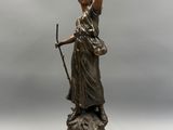 une statue antique en bronze représentant une femme tenant un bâton, posée sur un socle en bois