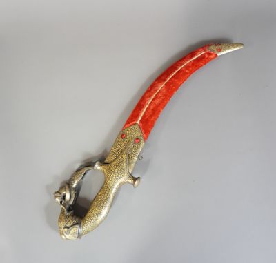 une image représentant un couteau en forme de lézard, de couleur rouge et or, posé sur une surface blanche