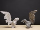 paire de figurines de coqs en métal argenté posées sur une table devant un fond noir