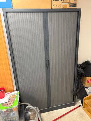une armoire métallique dans un coin de la pièce, avec des cartons et une serpillière posés par terre