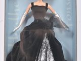La poupée se trouve dans une boîte, vêtue d'une robe noire et de gants blancs.