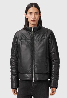 Un homme vêtu d'une veste en cuir noire et d'un jean se tient debout devant un fond blanc.