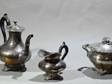 three silver teapots and two silver cups on a table