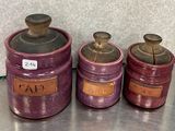 trois récipients en céramique violets, munis d'étiquettes, posés sur une table