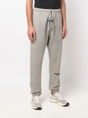Un homme vêtu d'un t-shirt noir et d'un pantalon de survêtement gris se tient devant un mur blanc.