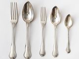 five silver spoons and a fork are lined up on a white surface