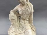 une statue blanche représentant une femme assise sur un rocher, avec une fleur dans les cheveux