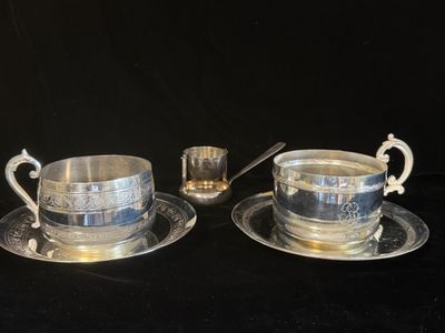 deux tasses et soucoupes en métal argenté, accompagnées de cuillères, posées sur un fond noir