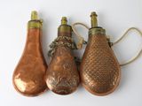 trois bouteilles de couleur cuivrée auxquelles sont suspendues des chaînes, posées sur une surface blanche
