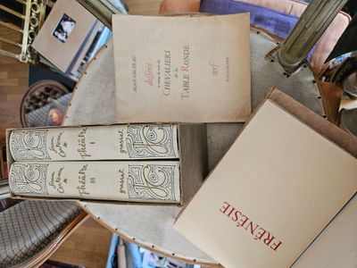 Plusieurs livres sont empilés sur une table, avec une chaise à l'arrière-plan.