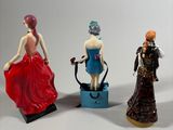 three figurines of women in dresses are posed on a white surface