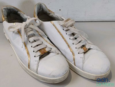 une paire de baskets blanches ornées de fermetures éclair dorées, posées sur une surface blanche